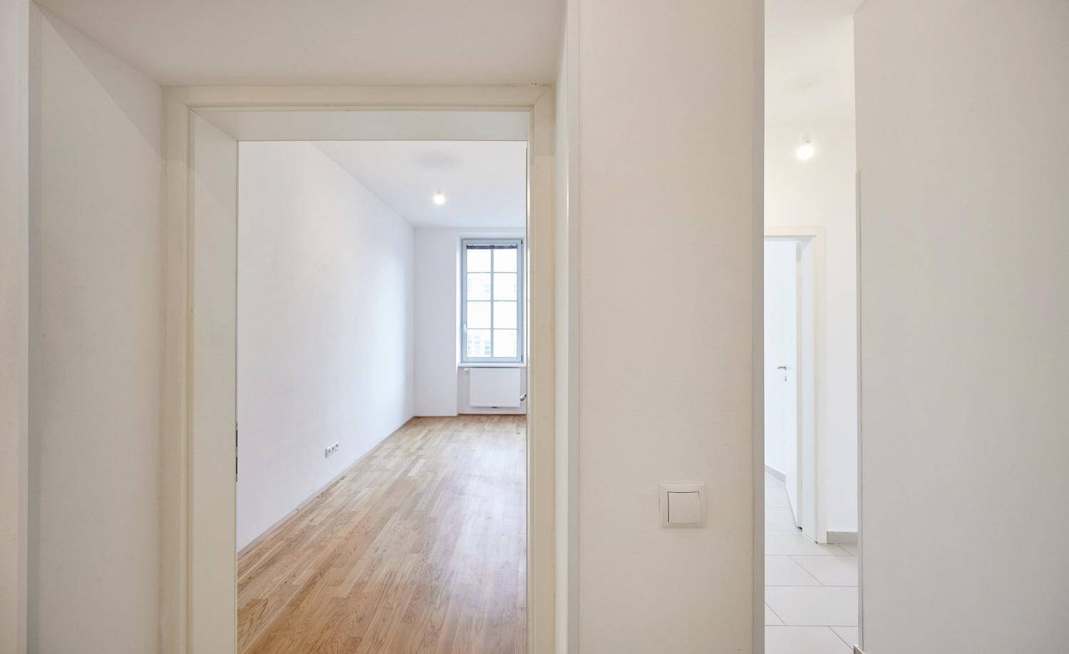 Heller Flur mit Zugang zu einem weiteren Zimmer mit Parkettboden und Fenster.