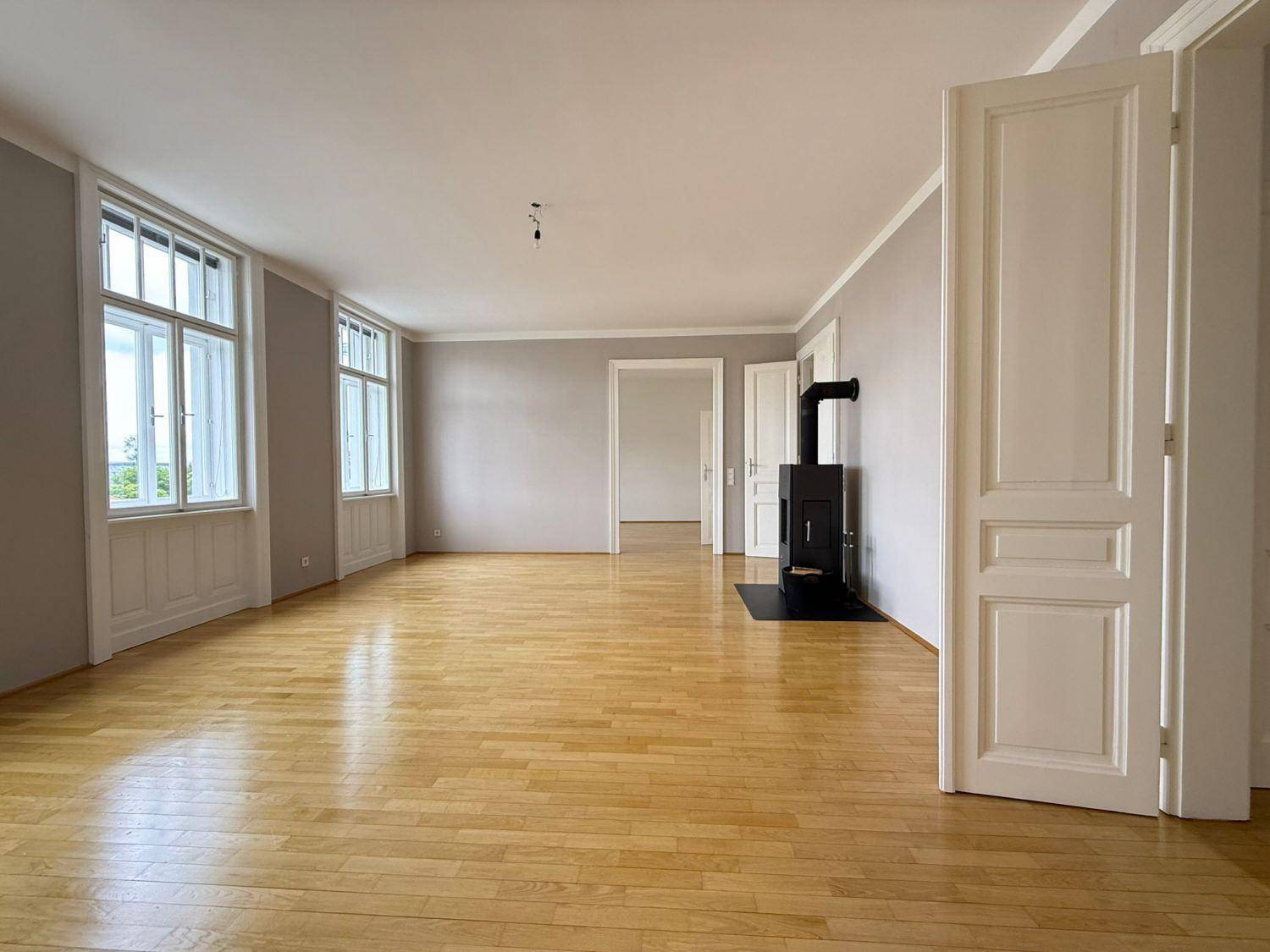 Geräumiges Wohnzimmer mit hellem Parkettboden, Kaminofen und viel Tageslicht durch hohe Fenster.