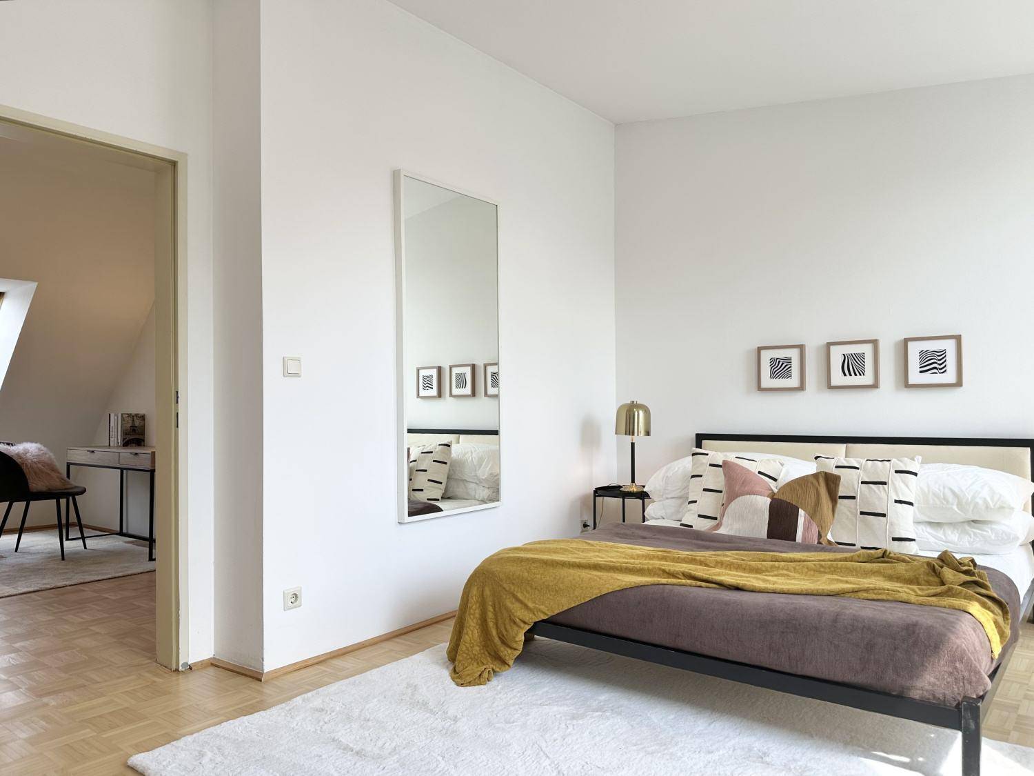 Schlafzimmer mit modernem Bett, Spiegel und Blick in den angrenzenden Arbeitsbereich.