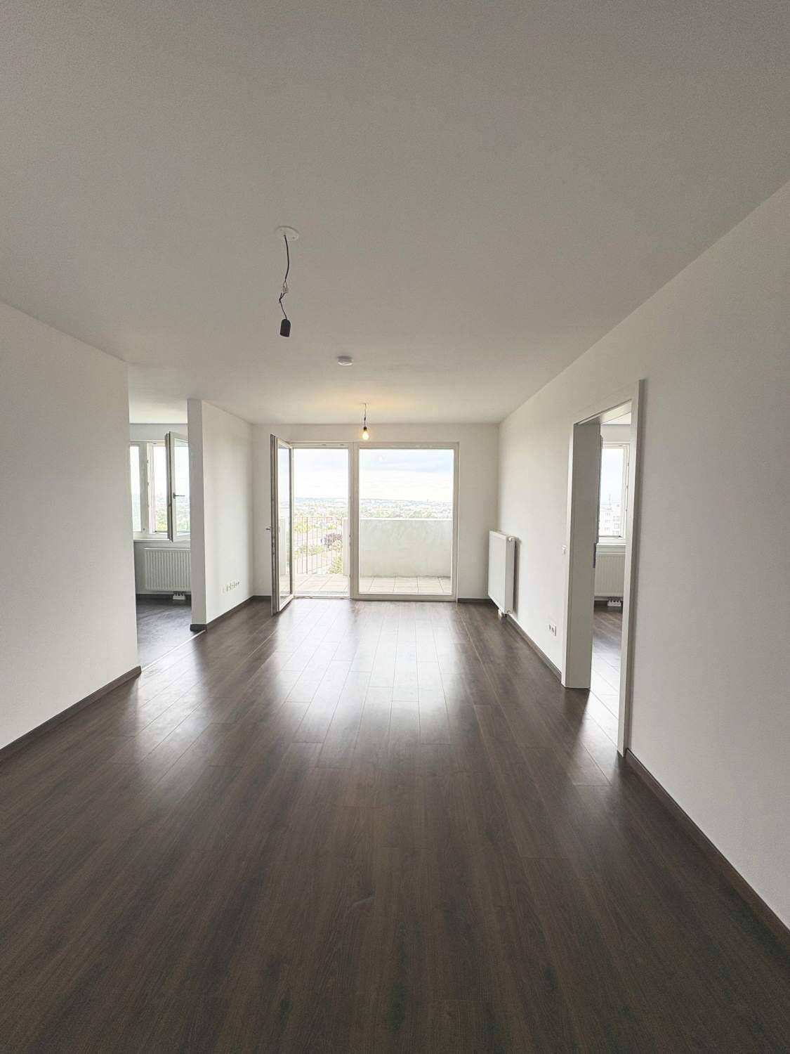 Helles Wohnzimmer mit dunklem Laminatboden, weißen Wänden und Zugang zum Balkon mit Weitblick.