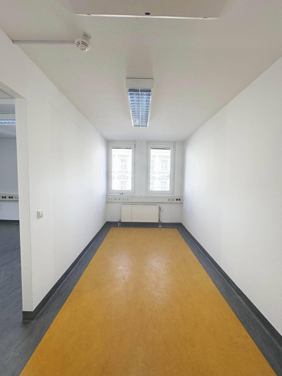 Kleinerer Büroraum oder Flurbereich mit auffälligem gelbem Boden und zwei Fenstern.