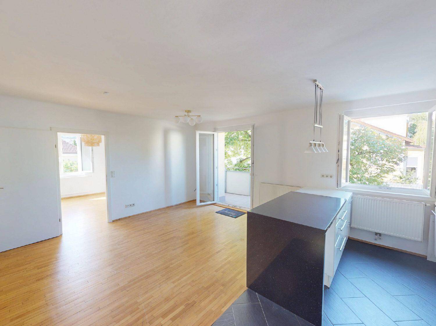 Geräumiger Wohnbereich mit Holzboden, offener Kücheninsel und hellem Balkonzugang.