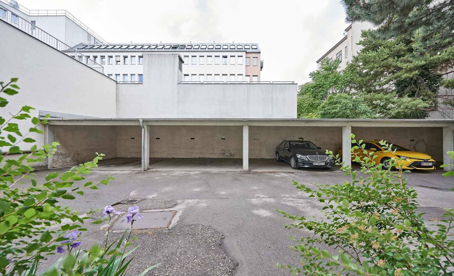 Außenansicht der überdachten Parkplätze mit zwei Fahrzeugen und grünen Büschen im Vordergrund.