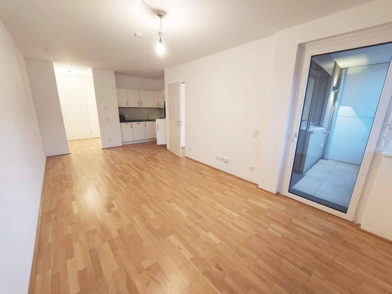 Geräumiger Wohnbereich mit Parkettboden, weißer Küchenzeile im Hintergrund und Balkonzugang.