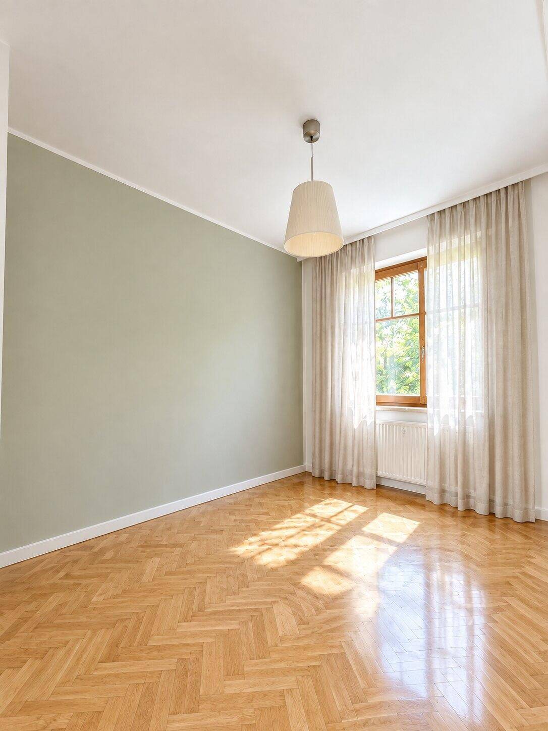 Helles Zimmer mit Fischgrätparkett, Fenster mit Vorhängen und viel Tageslicht.