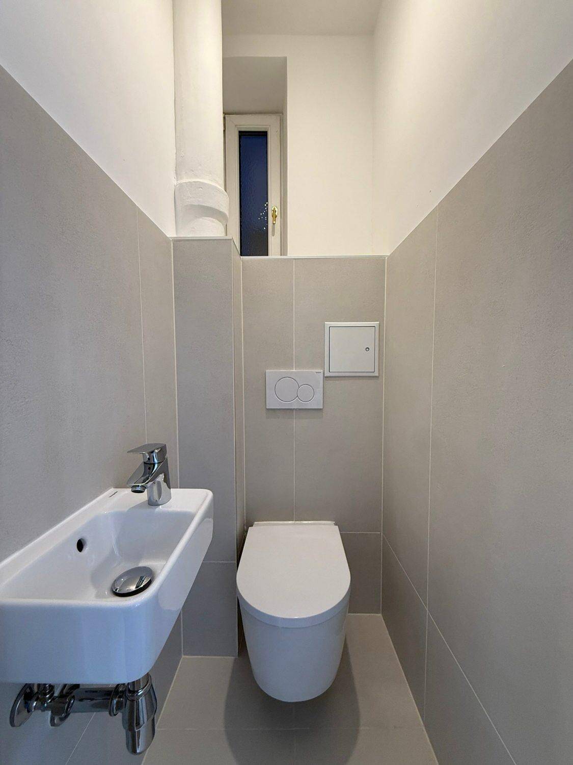 Kompaktes Gäste-WC mit wandhängender Toilette, kleinem Waschbecken und einem hohen Fenster.