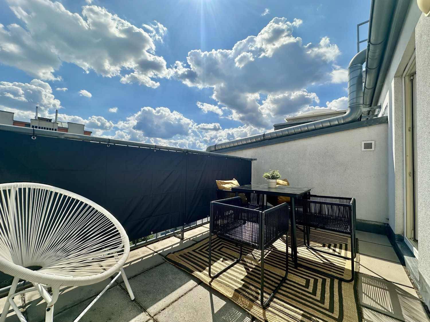 Sonniger Balkon mit Outdoor-Möbeln und Blick auf die Dächer der Stadt unter blauem Himmel.