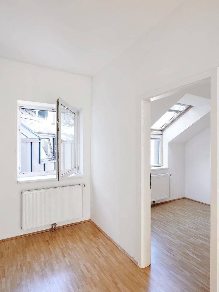 Helles Zimmer mit Fenster, Holzboden und Zugang zu einem weiteren Raum.
