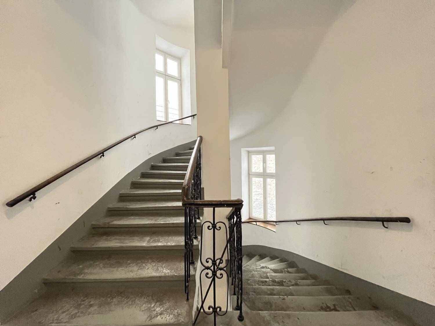 Geschwungenes Treppenhaus mit Steinstufen, schmiedeeisernem Geländer und Fenstern für Tageslicht.