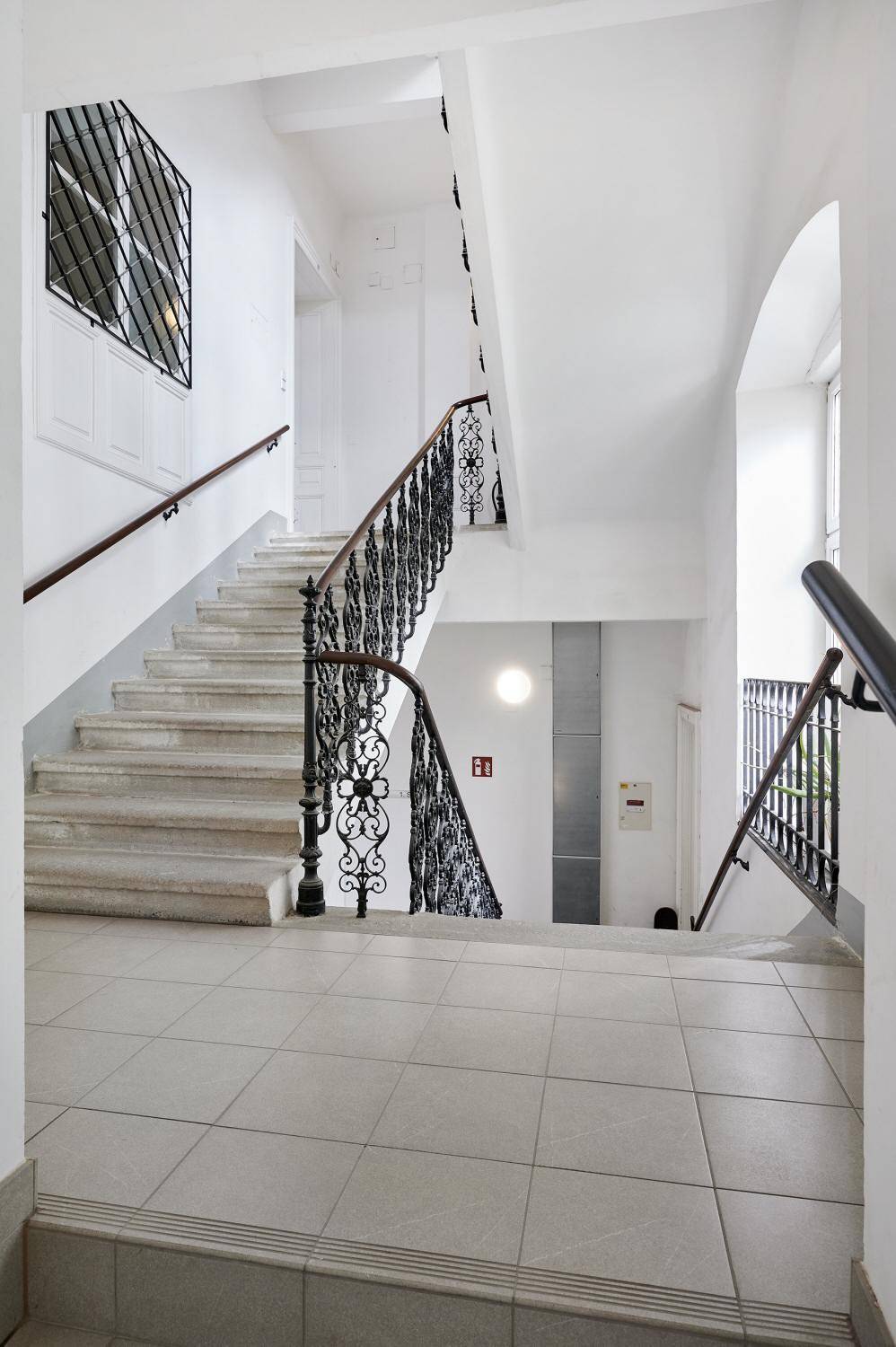 Gepflegtes Treppenhaus mit Steinstufen, schmiedeeisernem Geländer und hellem Fliesenboden.