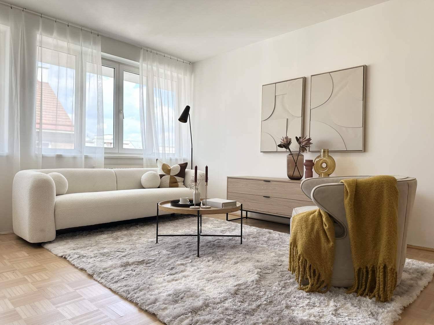 Gemütliches Wohnzimmer mit beiger Couch, flauschigem Teppich und großen Fenstern für viel Licht.