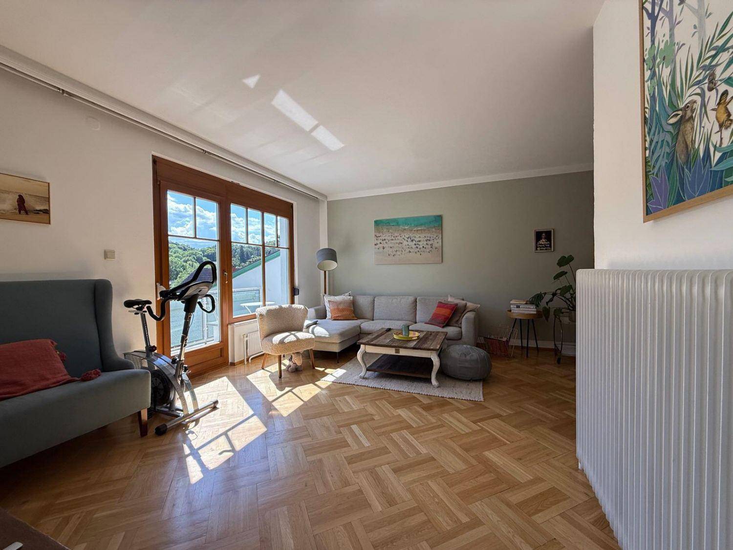 Helles Wohnzimmer mit Fischgrätparkett, großen Fenstern und Zugang zu einem Balkon.