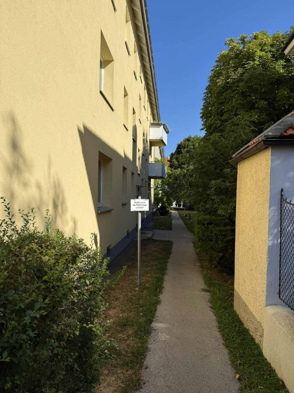 Seitenansicht des Gebäudes mit einem schmalen Gehweg und grüner Bepflanzung.