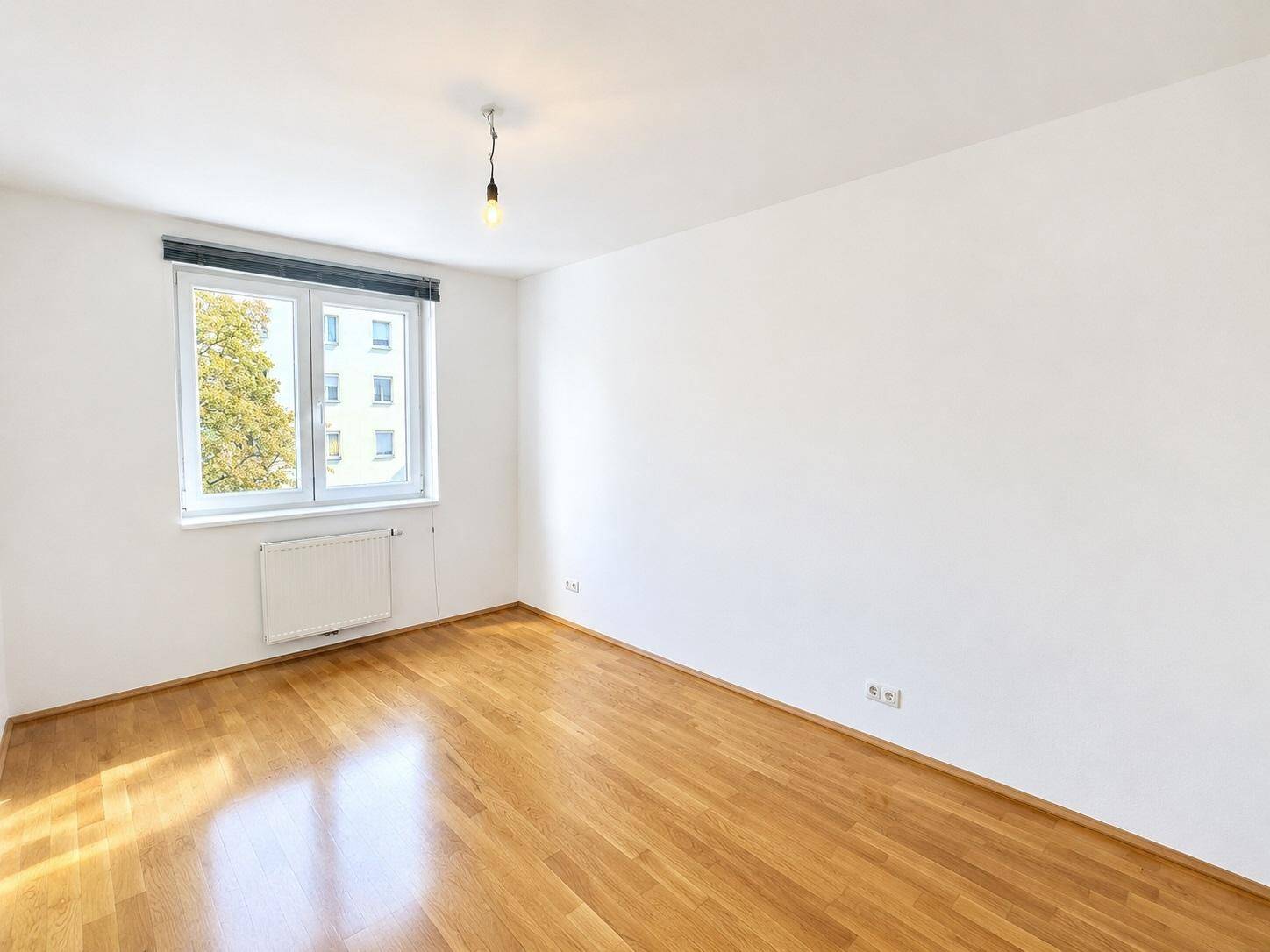 Leeres Schlafzimmer mit Parkettboden, weiß gestrichenen Wänden und Blick ins Grüne.