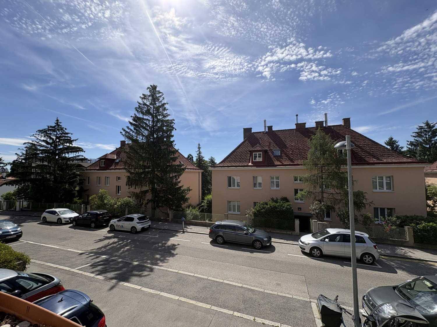 Blick auf die ruhige Wohnstraße mit parkenden Autos und umliegenden Gebäuden.