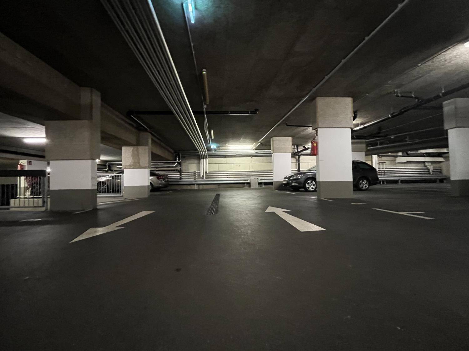 Geräumige Tiefgarage mit Betonpfeilern, sichtbaren Rohren und Richtungspfeilen auf dem dunklen Boden.