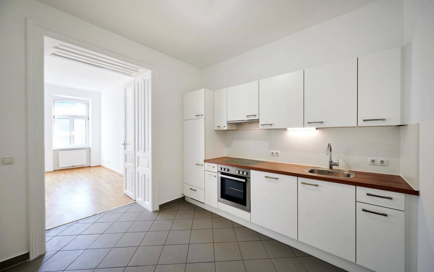 Funktionale Küche mit weißen Schränken, Holzarbeitsplatte und Fliesenboden, angrenzend an ein weiteres Zimmer.