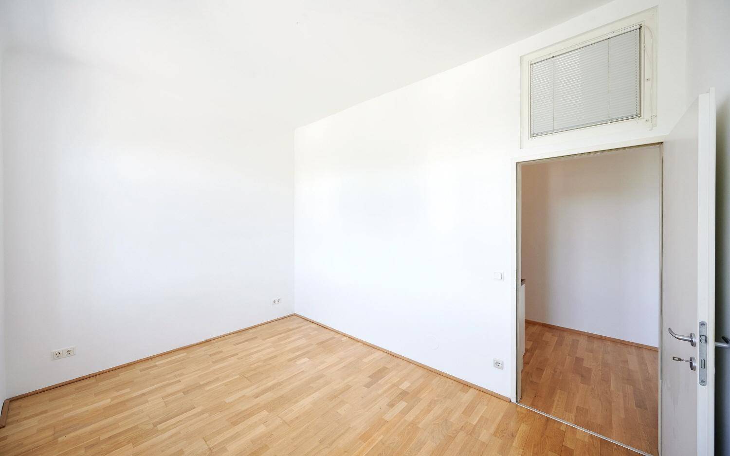 Helles, leeres Zimmer mit Parkettboden und einem Fenster über der Tür.