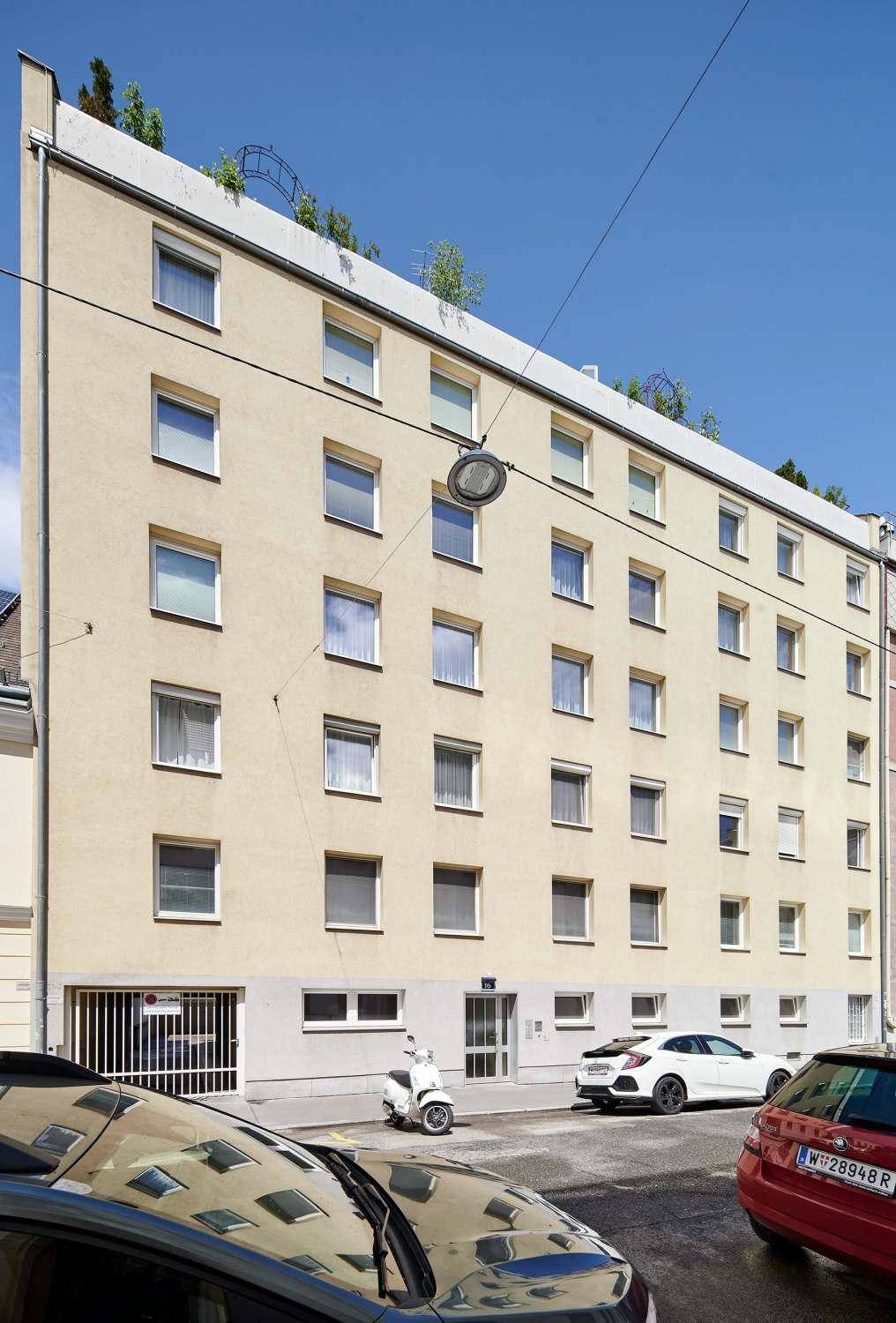 Schrägansicht der hellen Fassade eines Mehrfamilienhauses mit vielen Fenstern und klarem Himmel.