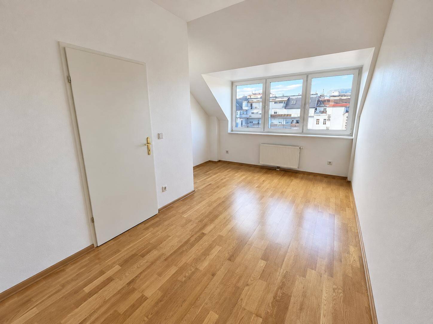 Helles Zimmer im Dachgeschoss mit Holzfußboden, schrägen Wänden und Blick auf die Stadt.