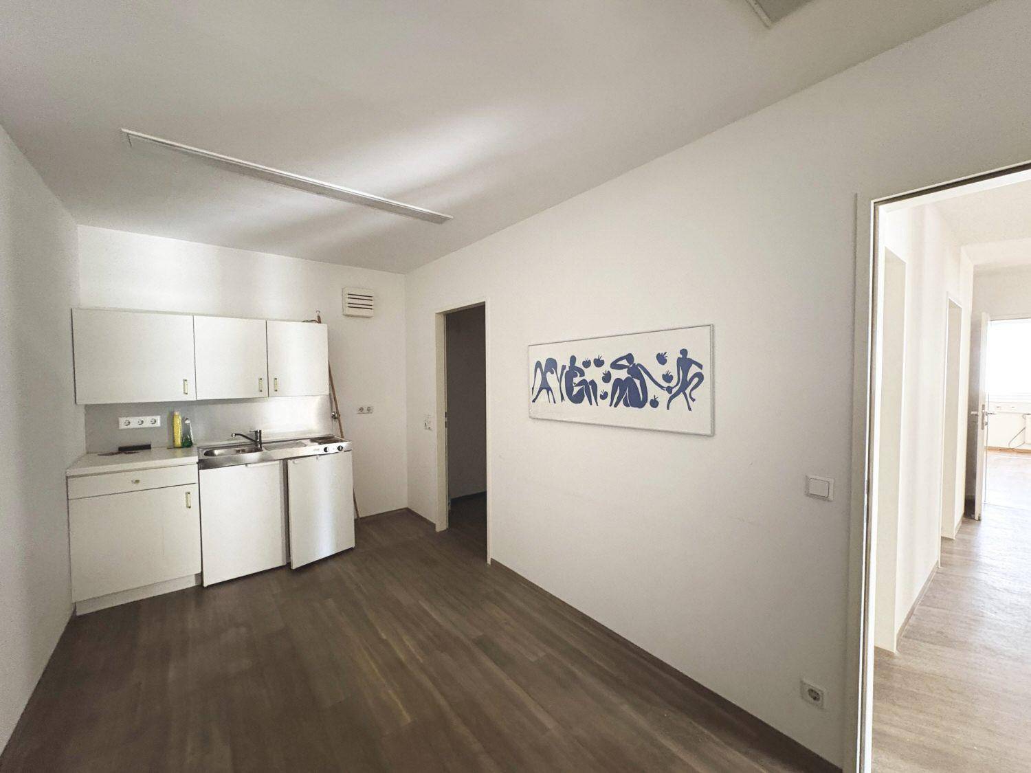 Einfache Küchenzeile mit weißen Schränken, Spüle und Kühlschrank in einem hellen Raum.