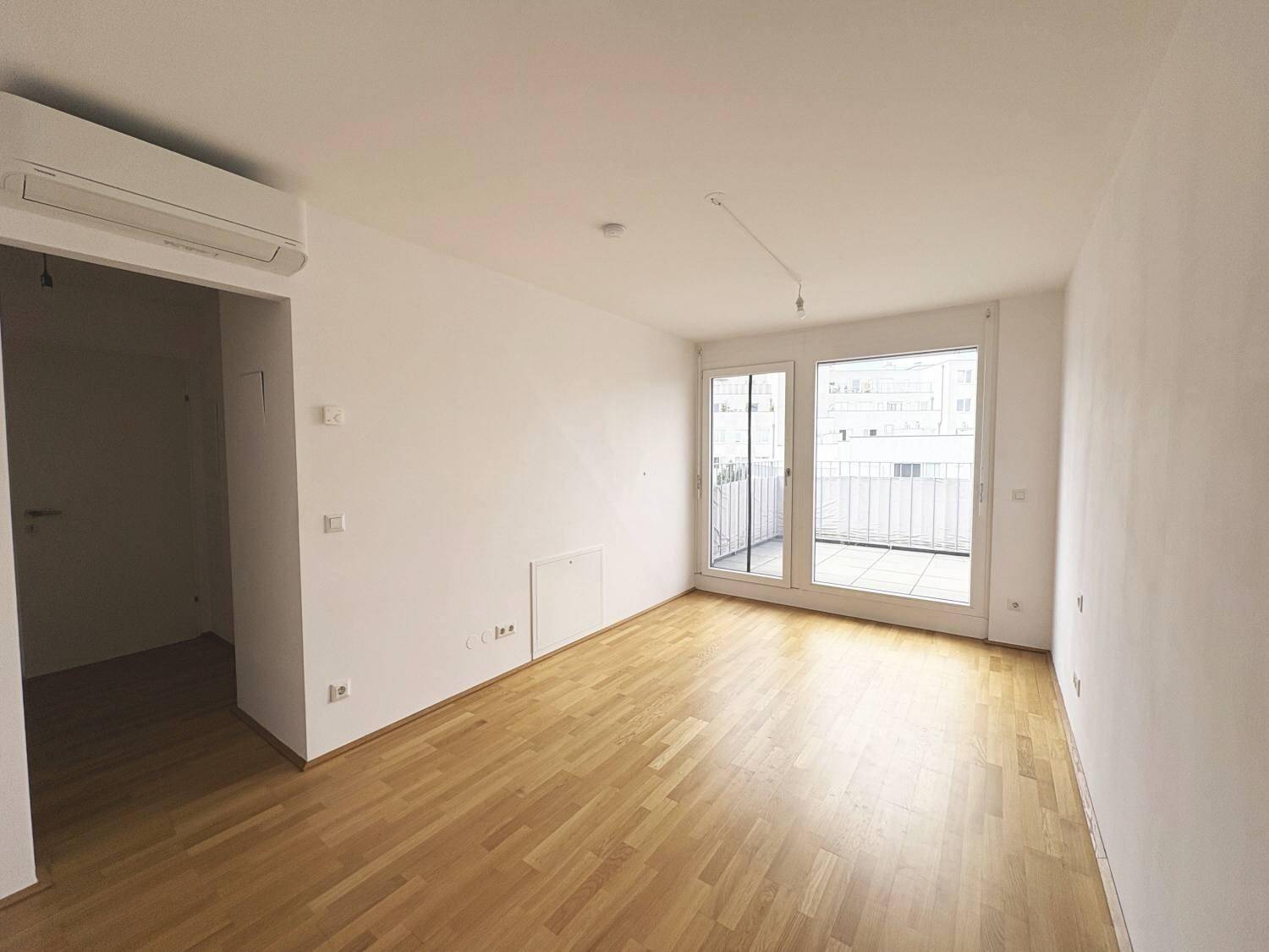 Heller Wohnraum mit Holzboden, weißen Wänden und großen Fenstern mit Zugang zu einem Außenbereich.