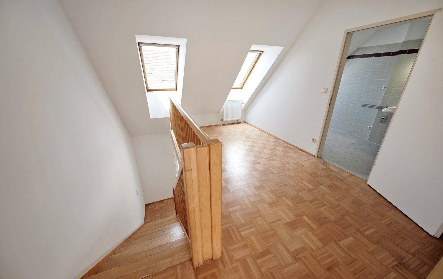 Dachgeschossraum mit schrägen Wänden, Parkettboden und zwei Dachfenstern, die den Raum erhellen.