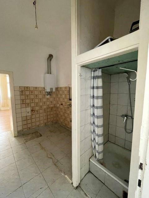 Einfaches Badezimmer mit Duschbereich, Vorhang und Warmwasserbereiter, in originalem Zustand.