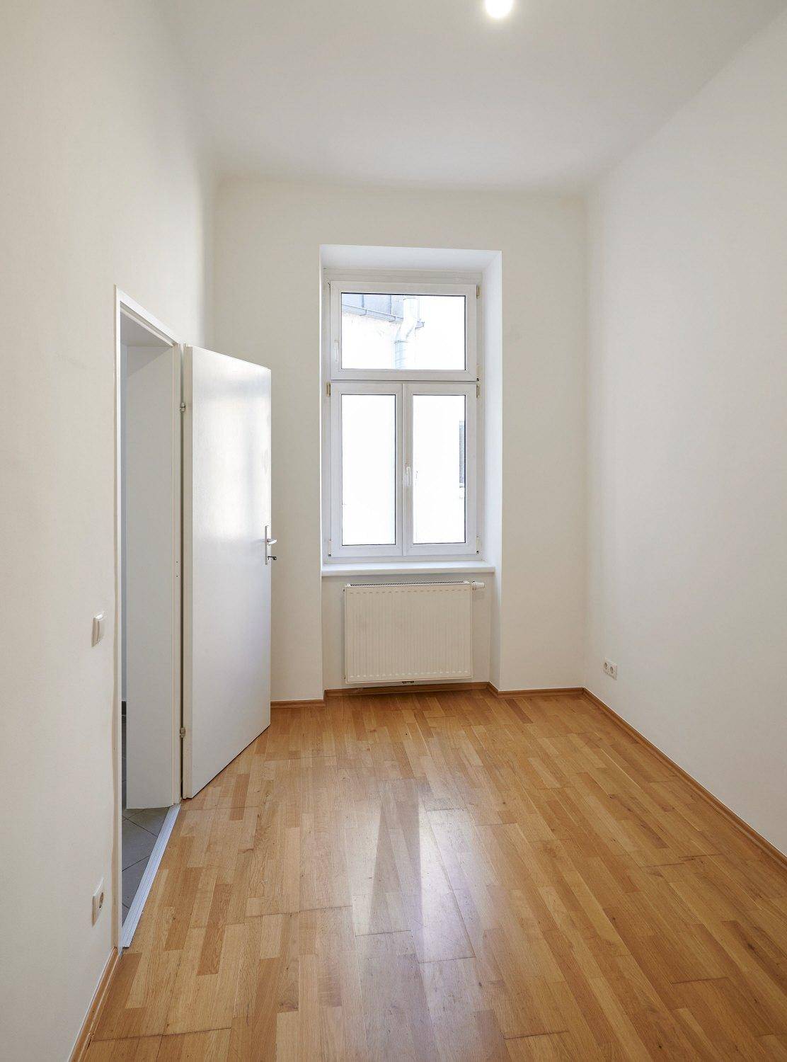 Helles, leeres Zimmer mit Parkettboden und einem Fenster, ideal als Schlaf- oder Arbeitszimmer.