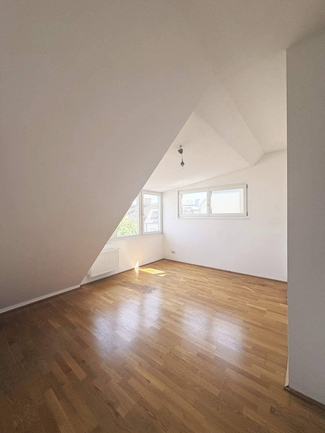 Lichtdurchflutetes Zimmer mit Holzboden, Dachschräge und Fenstern, die viel Sonnenlicht hereinlassen.