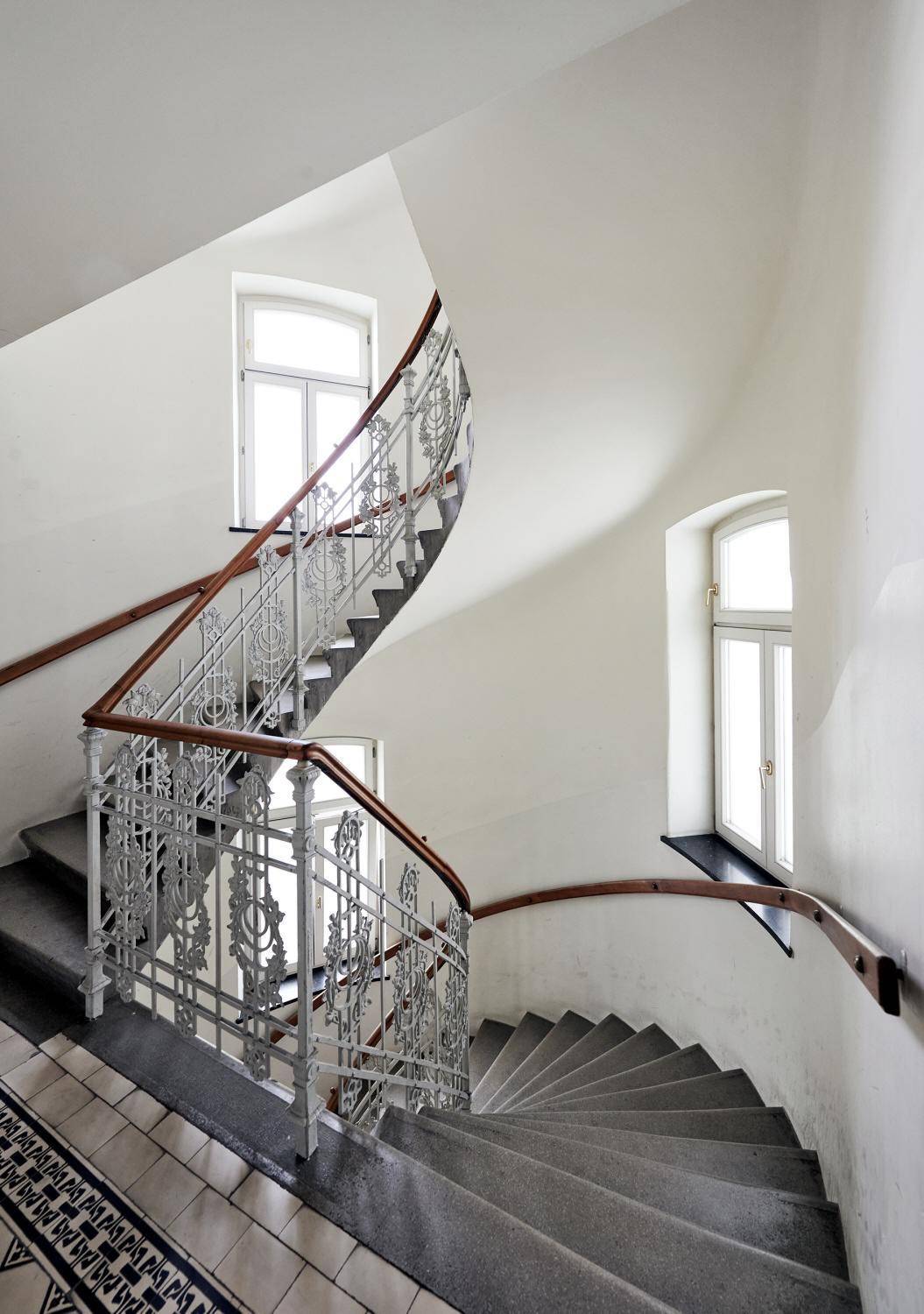 Elegantes Treppenhaus mit geschwungenem Geländer und großen Fenstern für Tageslicht.