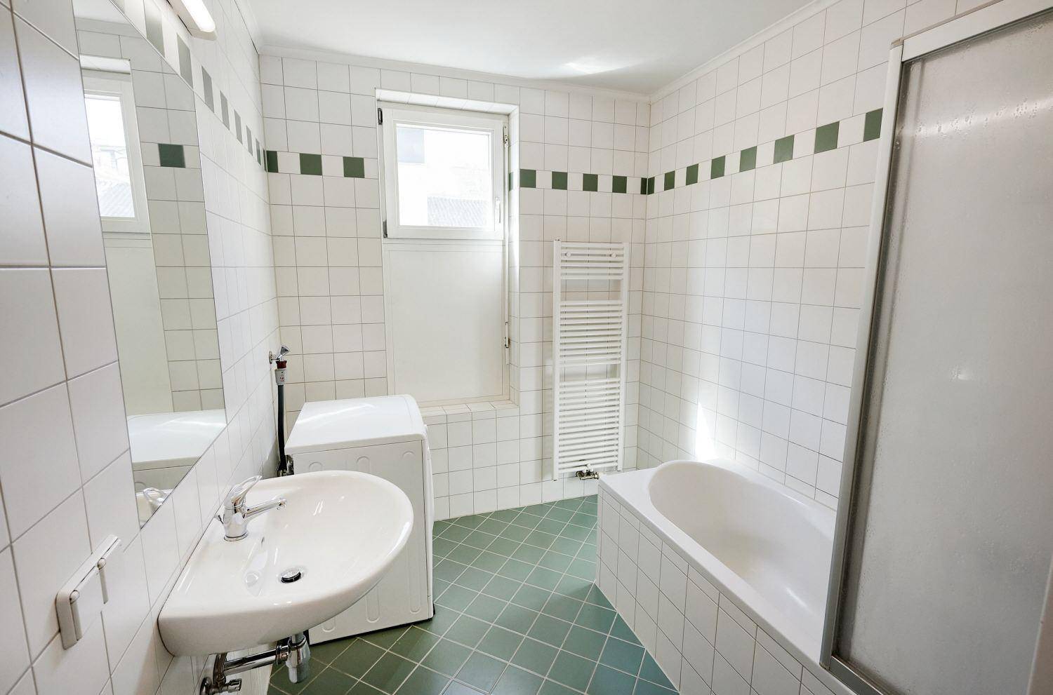 Badezimmer mit Badewanne, Waschbecken, Fenster und grün-weißen Fliesen.
