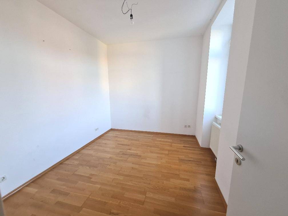 Helles, leeres Schlafzimmer mit Parkettboden und einem Fenster.