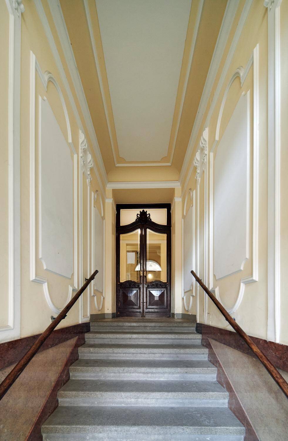 Elegante Eingangshalle mit breiter Steintreppe, verzierten Wänden und einer großen Holztür am Ende.