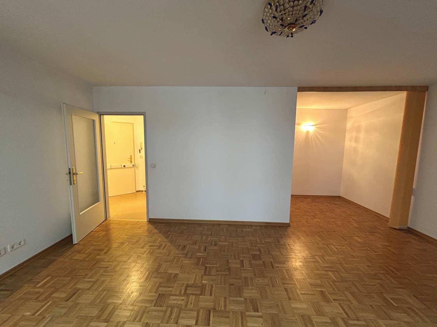 Geräumiges Wohnzimmer mit Parkettboden, Blick auf den Eingangsbereich und einen angrenzenden Nischenraum.