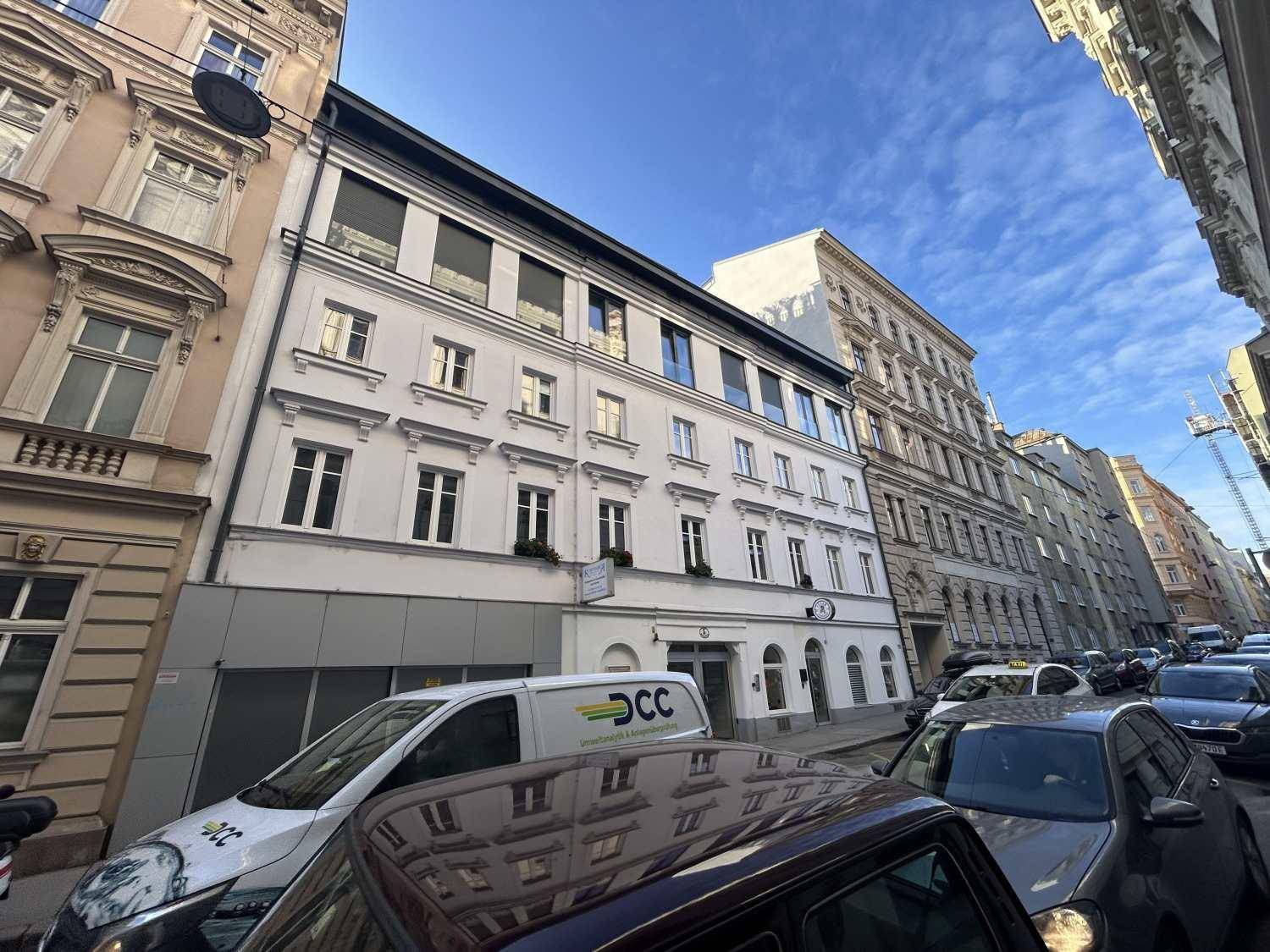 Weißes Gebäude mit klassischer Fassade an einer belebten Stadtstraße mit geparkten Autos.