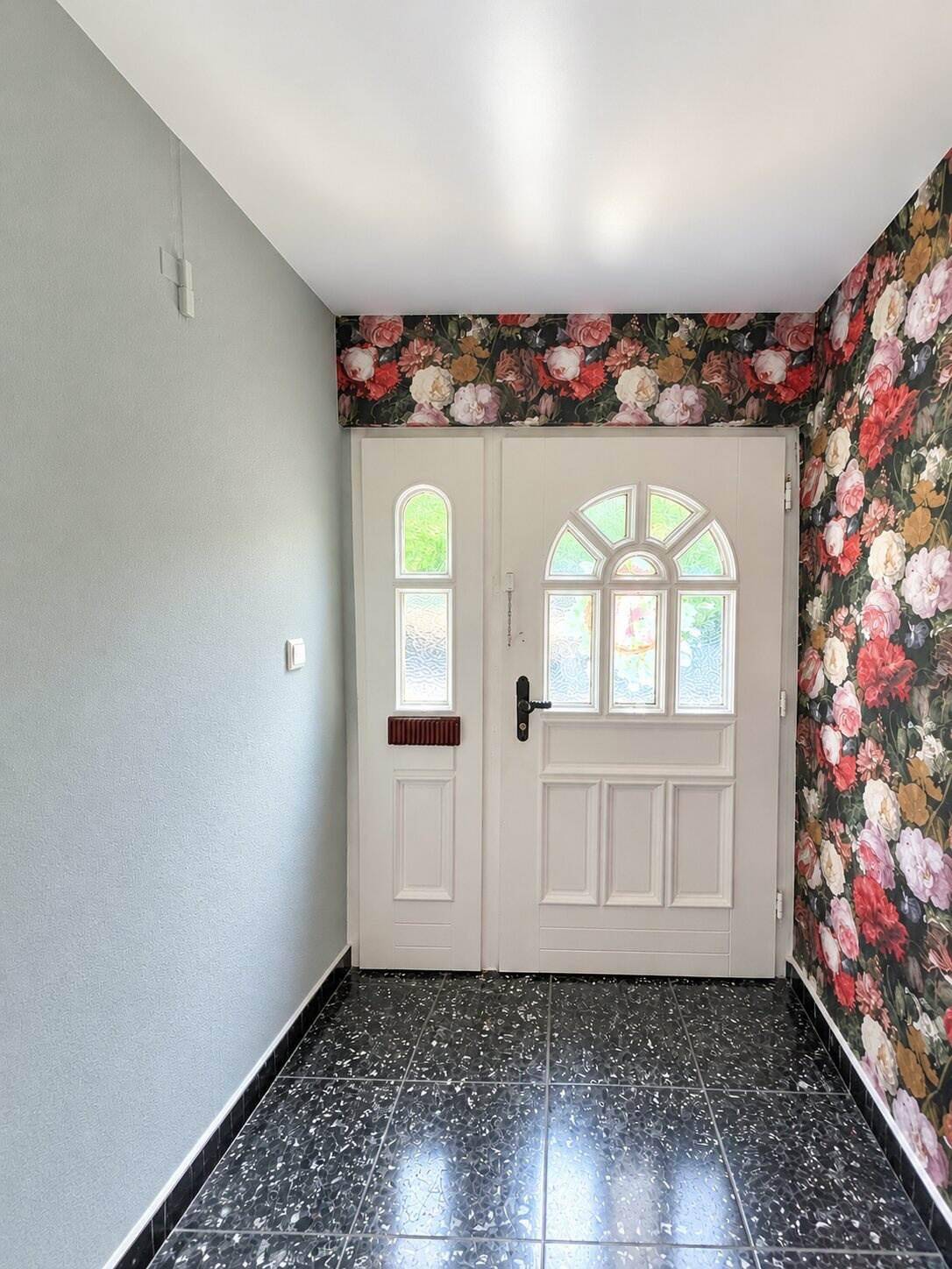 Einladender Eingangsbereich mit floraler Tapete, doppelter Eingangstür und dunklem Fliesenboden.
