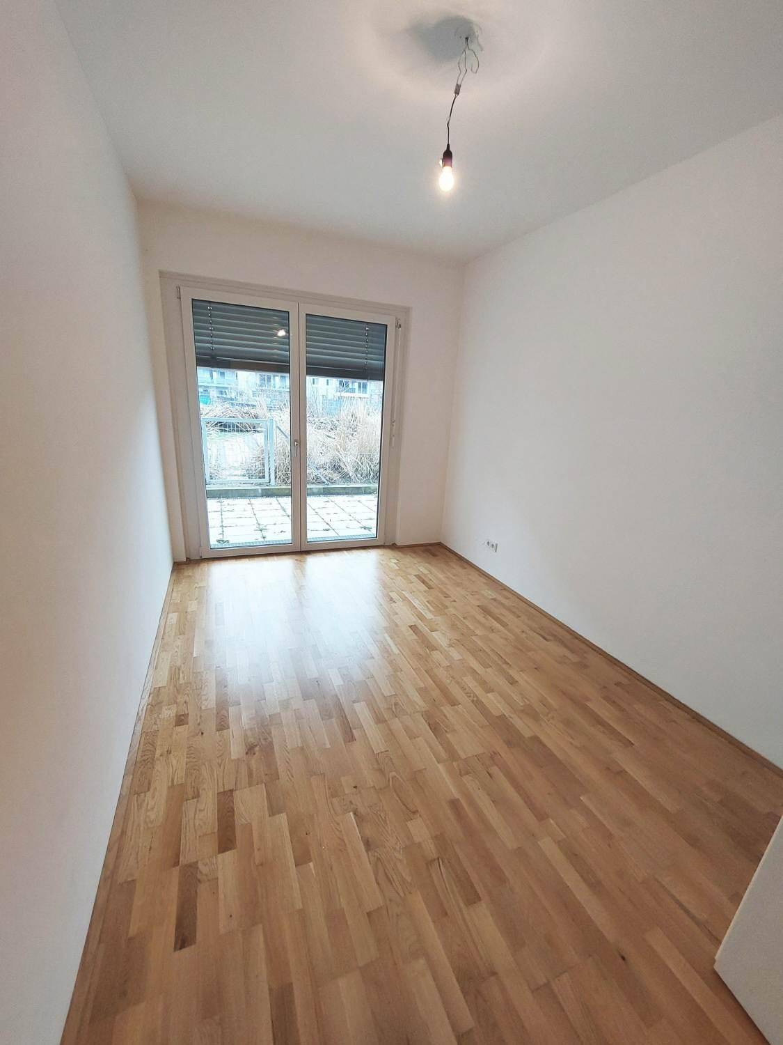 Helles Zimmer mit Parkettboden und großem Fenster mit Rollläden und Terrassenzugang.