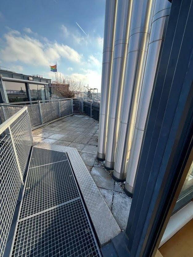 Teil der Dachterrasse mit Metallgeländer, großen Rohren und Blick auf den blauen Himmel.