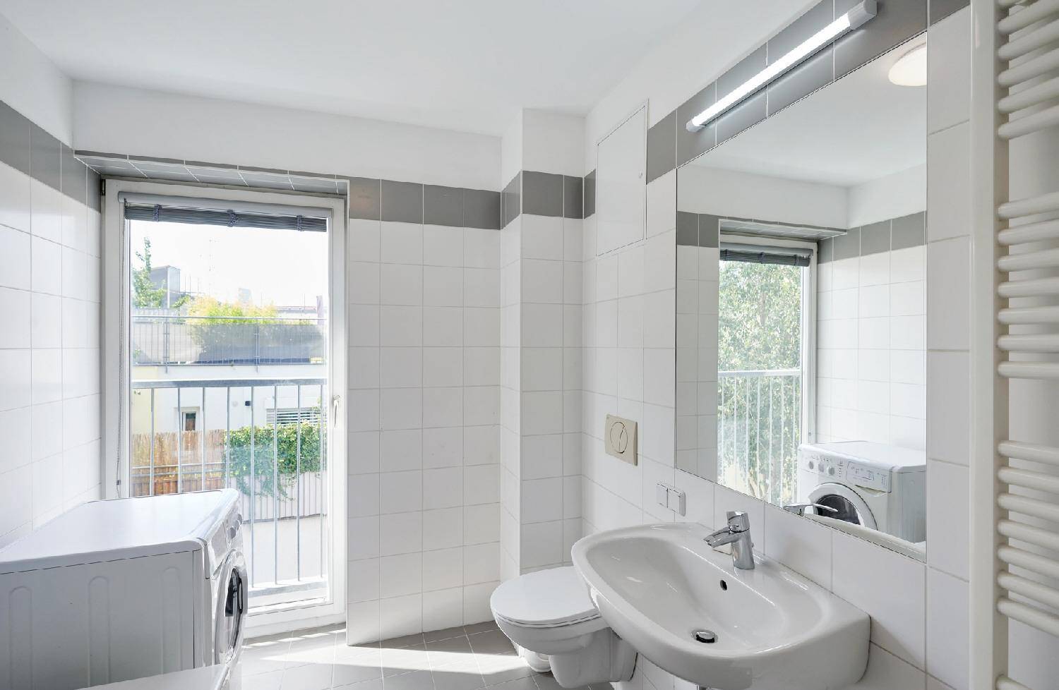 Helles Badezimmer mit Waschbecken, WC, Spiegel und Waschmaschine, mit Blick nach draußen.