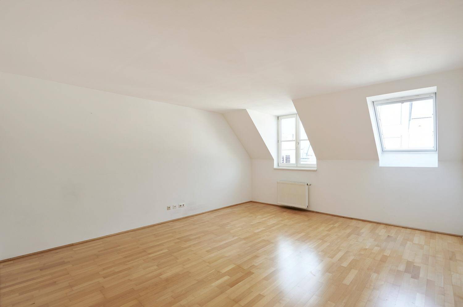 Geräumiger, heller Raum mit Holzparkett, Dachschrägen und zwei Fenstern, ideal als Wohn- oder Schlafzimmer.