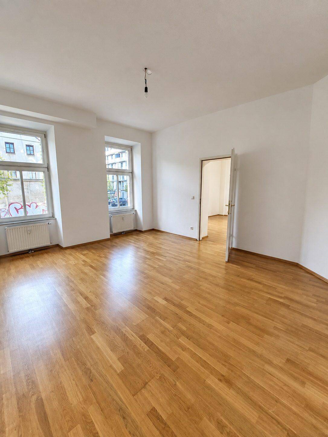 Großes, leeres Wohnzimmer mit glänzendem Parkettboden und zwei Fenstern, die den Raum erhellen.