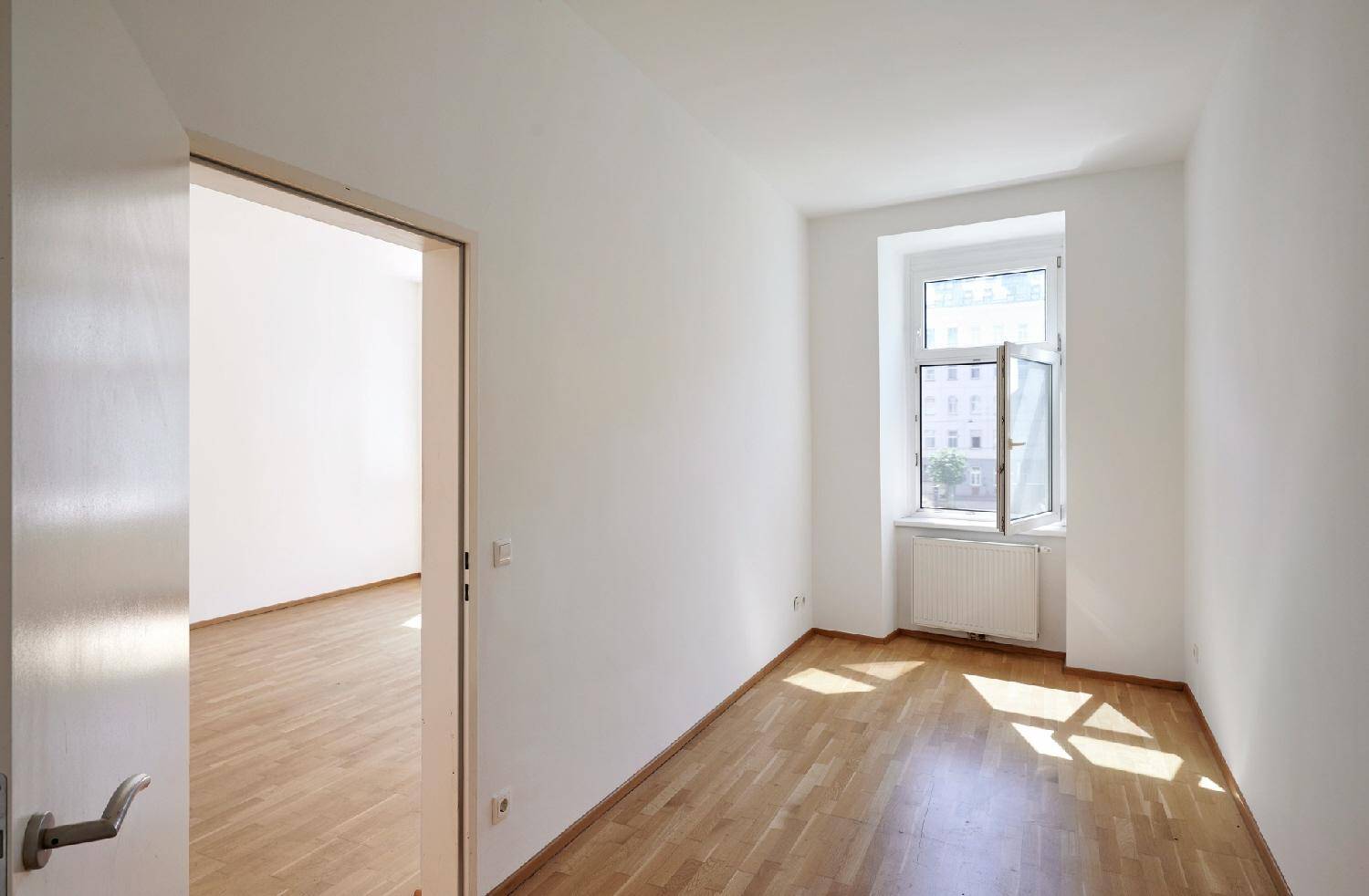 Helles Zimmer mit Parkettboden und Fenster, das Sonnenlicht hereinlässt, mit Zugang zu einem weiteren Raum.