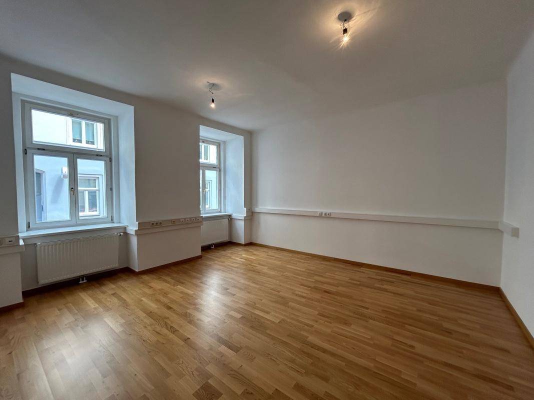 Helles Wohnzimmer mit zwei Fenstern, Holzboden und weißen Wänden.