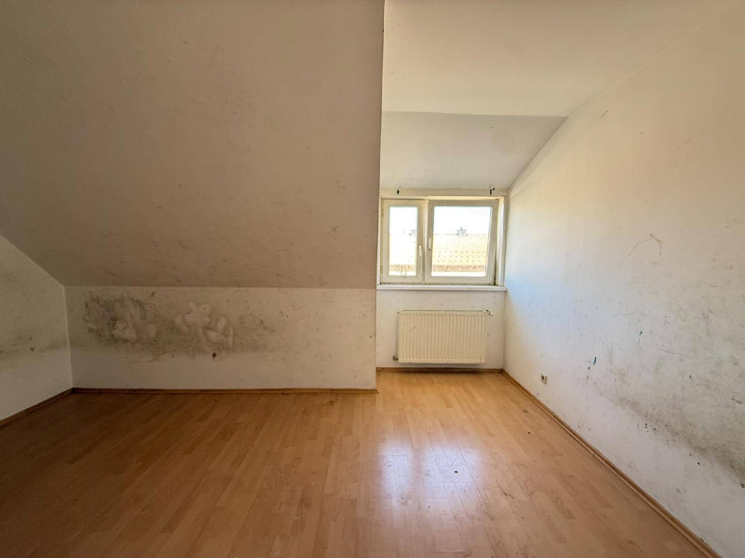 Dachgeschosszimmer mit schrägen Wänden, Fenster und Heizkörper, renovierungsbedürftig.