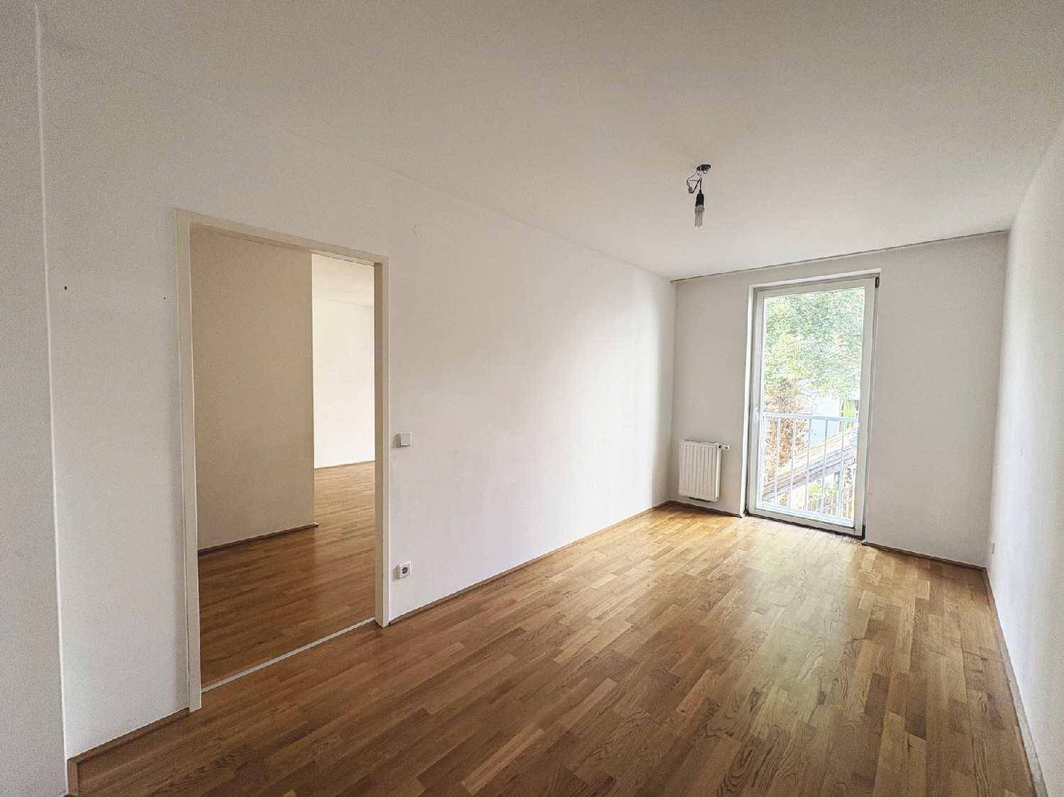 Leerer Raum mit Parkettboden und Zugang zu einem kleinen Balkon, der Tageslicht hereinlässt.