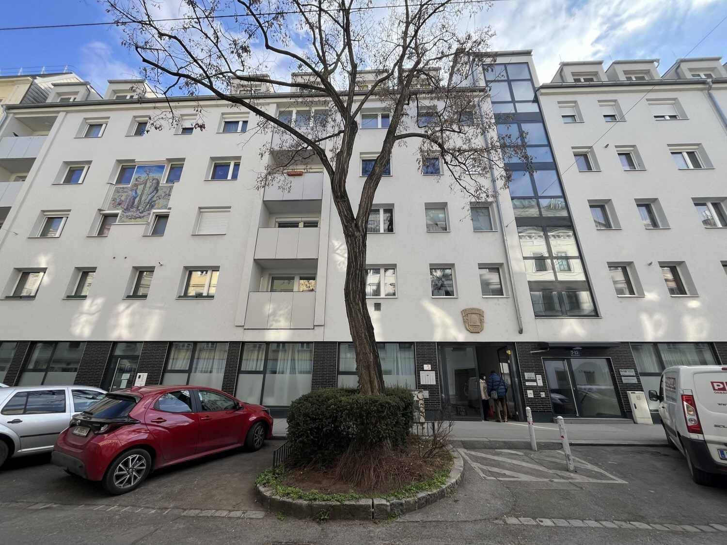 Mehrstöckiges Wohngebäude mit moderner Glasfassade und geparkten Autos davor in urbaner Umgebung.