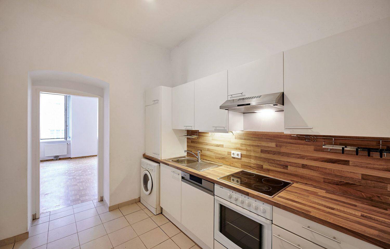 Moderne Küche mit weißen Fronten, Holzarbeitsplatte und Zugang zu einem weiteren Zimmer.