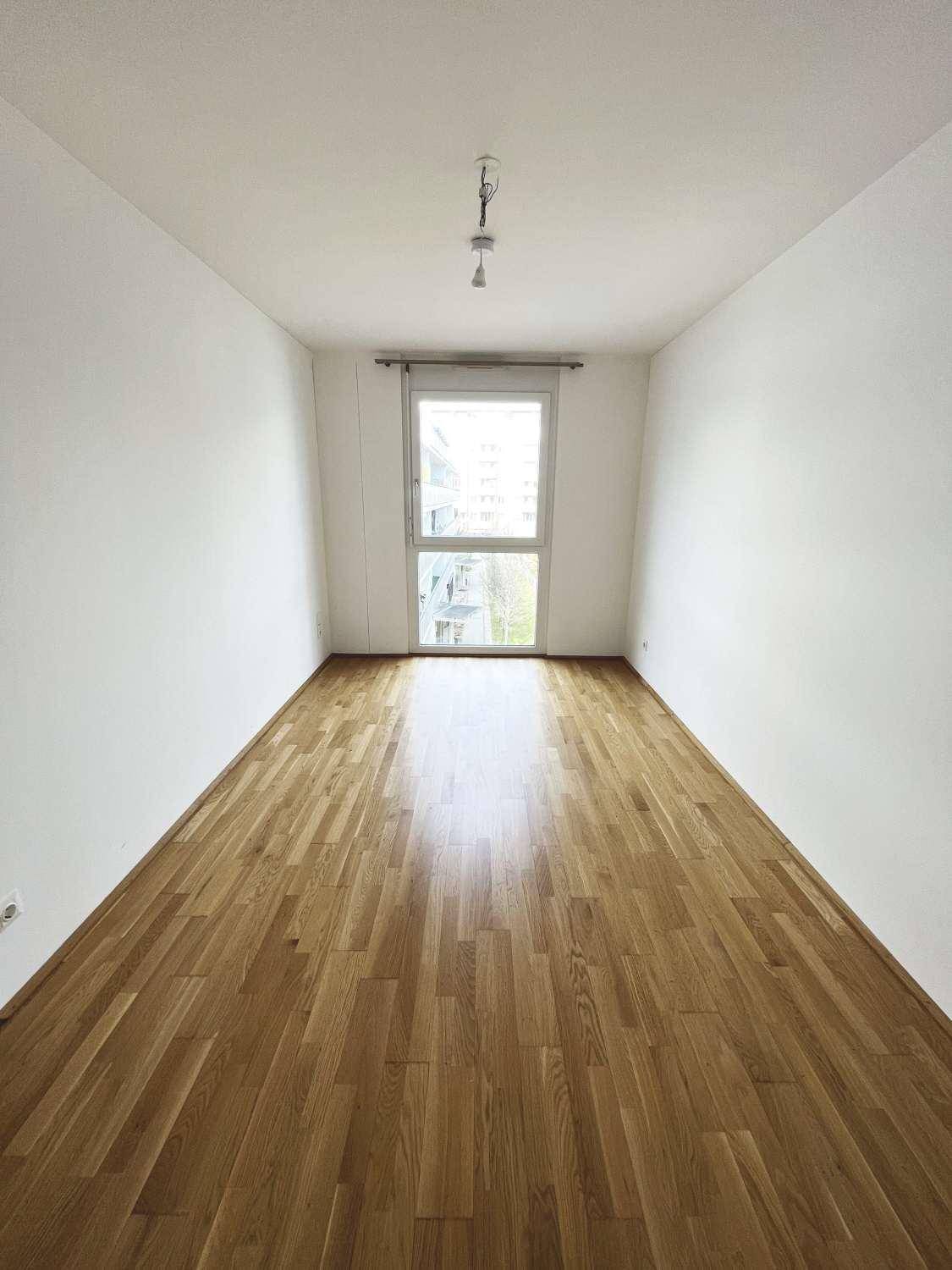 Langes, helles Zimmer mit Parkettboden und einem großen Fenster, das viel Tageslicht hereinlässt.