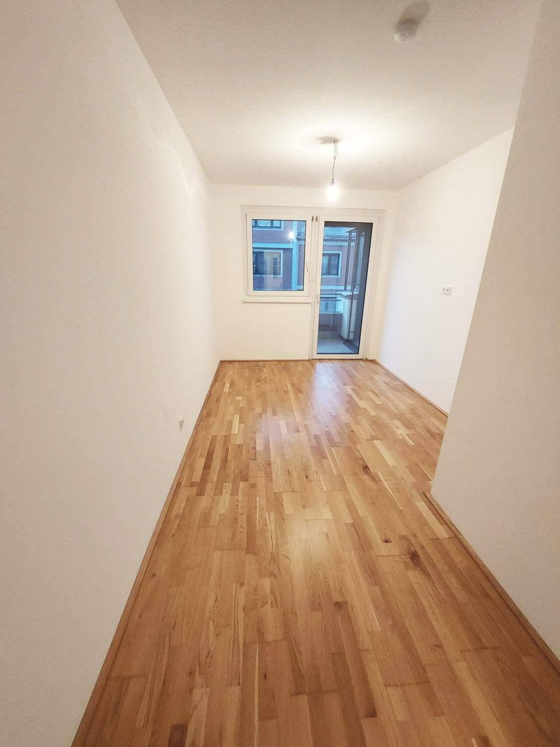 Heller, leerer Wohnraum mit Parkettboden und Fensterfront, die viel Tageslicht hereinlässt.